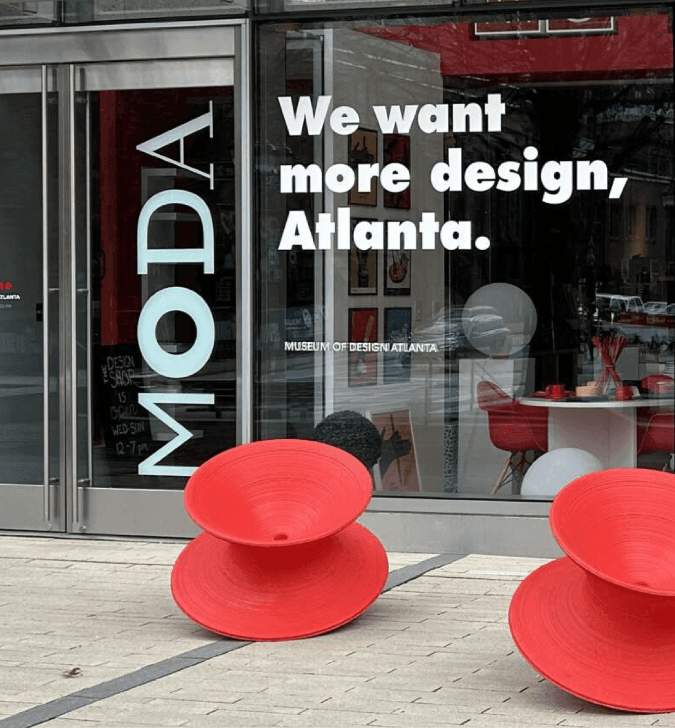 The MODA storefront features large white window text reading “We want more design, Atlanta” beside red spinning chairs outside the museum.