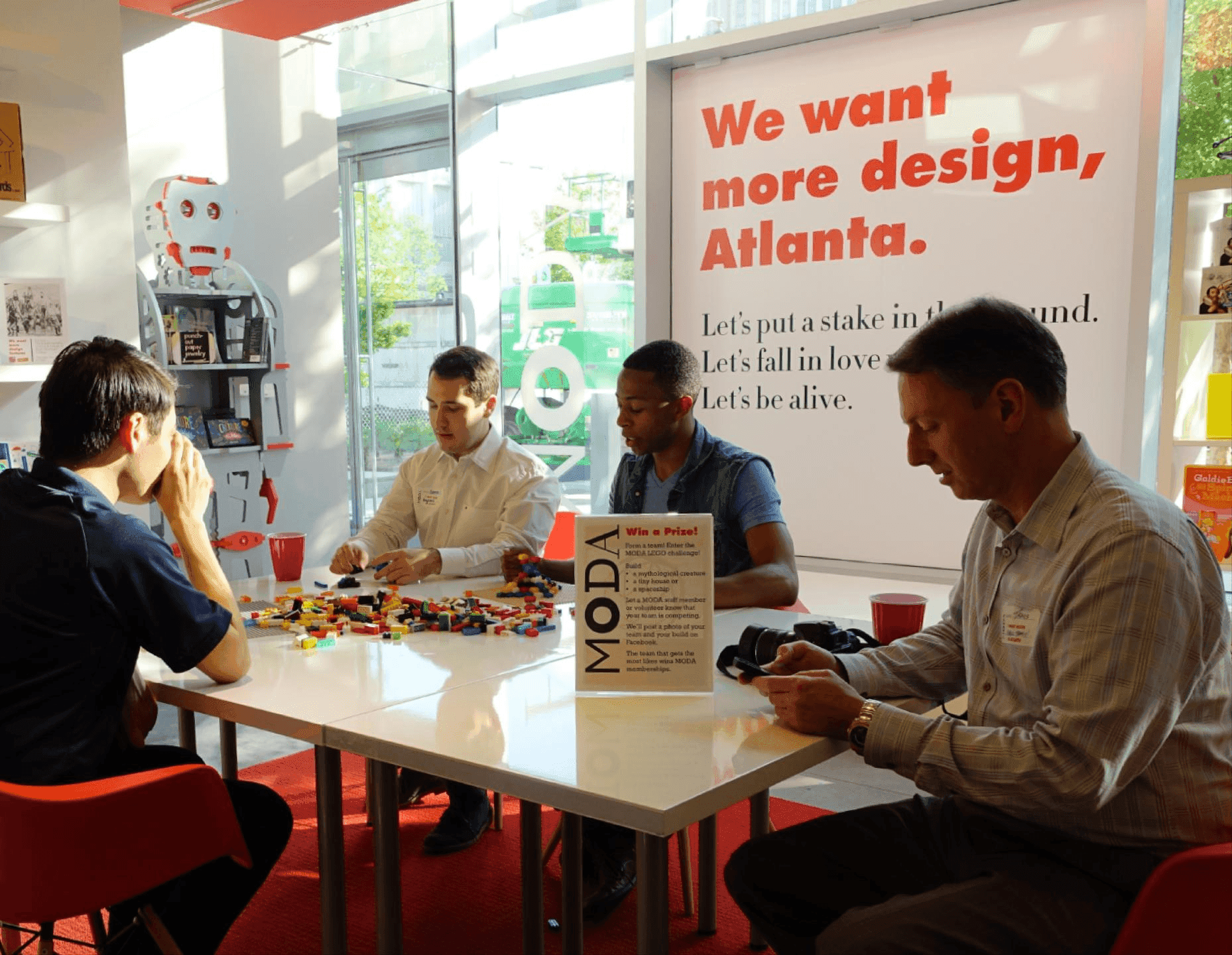 Visitors gather around a table building with LEGO bricks inside MODA, with a large wall graphic behind them reading “We want more design, Atlanta.”